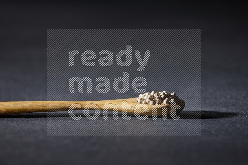 A wooden spoon full of white pepper beads on black flooring
