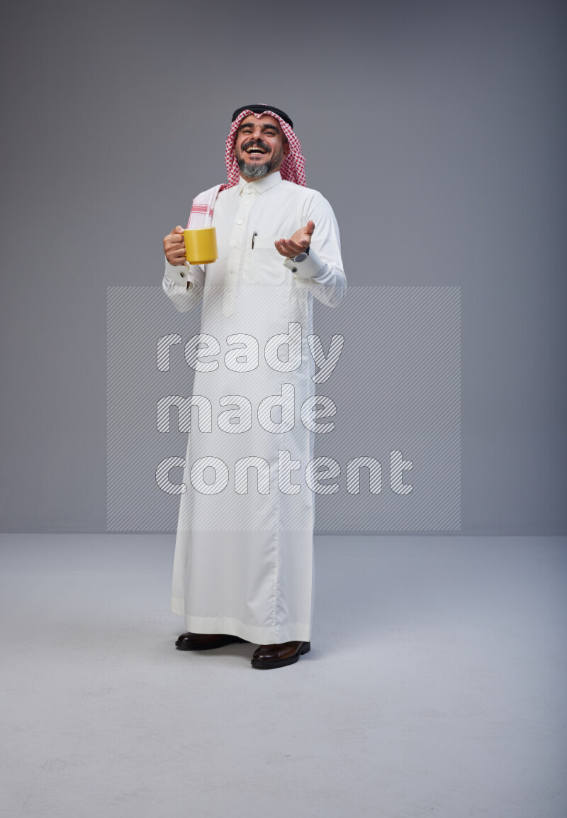 Saudi man Wearing Thob and red Shomag standing holding mug on Gray background