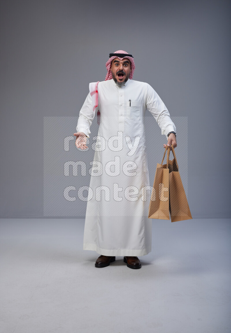 Saudi man Wearing Thob and red Shomag standing holding shopping bag on Gray background