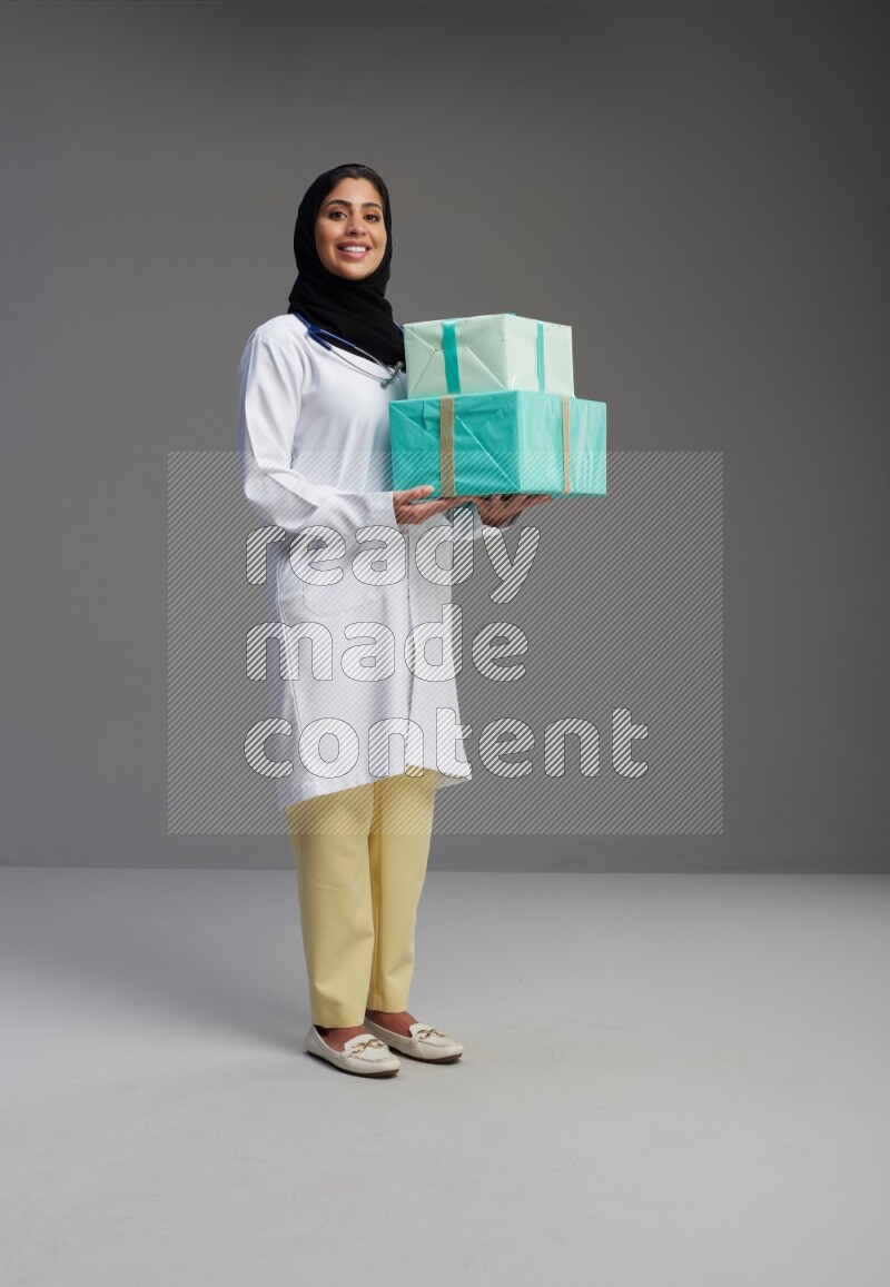 Saudi woman wearing lab coat with stethoscope standing holding gift box on Gray background