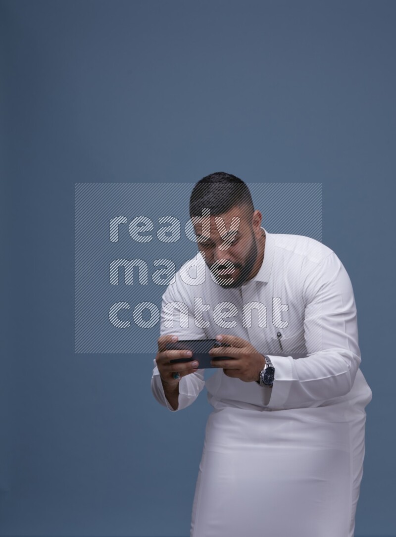 A man Playing a Game on his Smartphone  on Blue Background wearing Saudi Thob