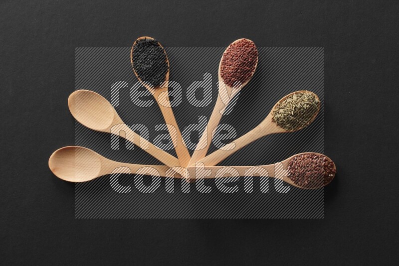 wooden spoons filled with white peppers, mustard seeds, black seeds, garden cress, cumin and flax on black flooring