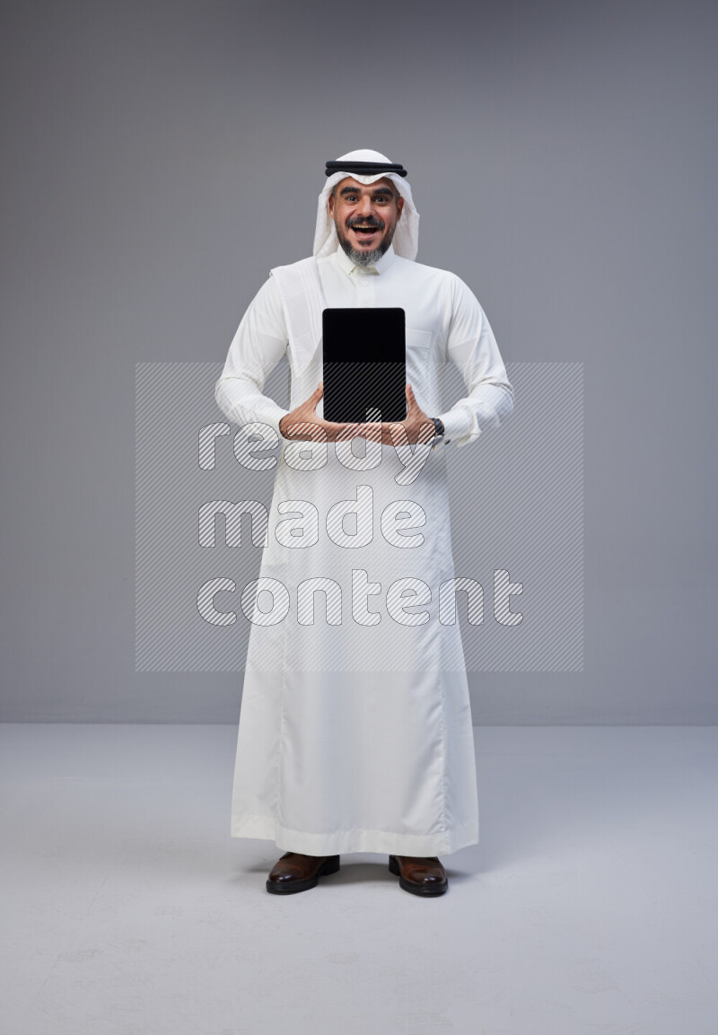 Saudi man Wearing Thob and white Shomag standing showing tablet to camera on Gray background