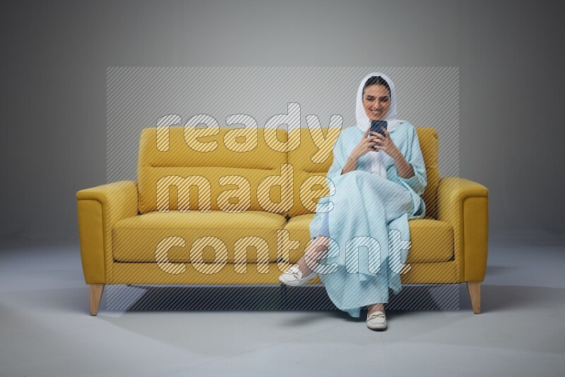 A Saudi woman wearing a light blue Abaya and white head scarf sitting on a yellow sofa and using her phone eye level on a grey background