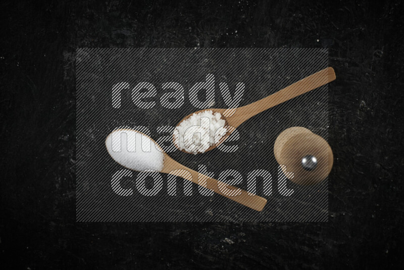 2 wooden spoons filled with fine and coarse salt with wooden grinder beside them on black background