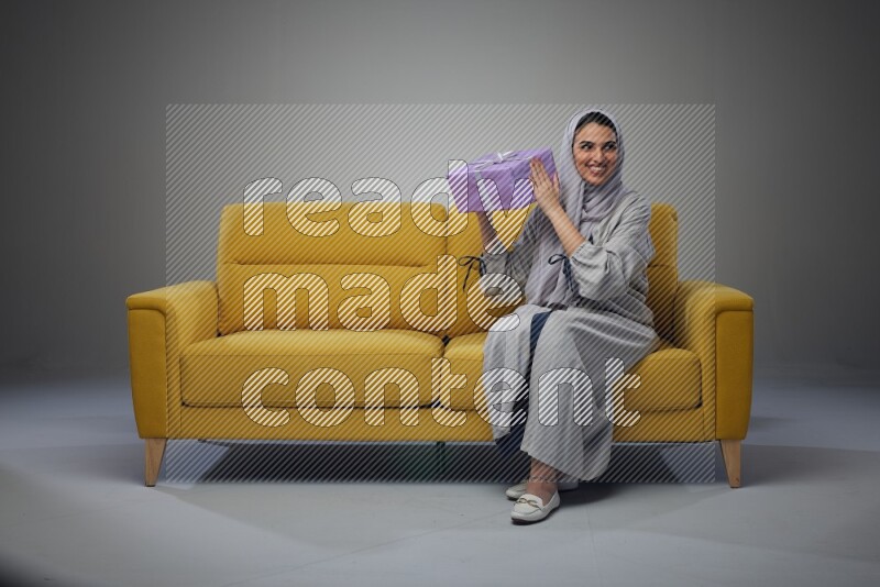 A Saudi woman wearing a light gray Abaya and head scarf sitting on a yellow sofa holding a gift box beside multi color gift boxes eye level on a grey background