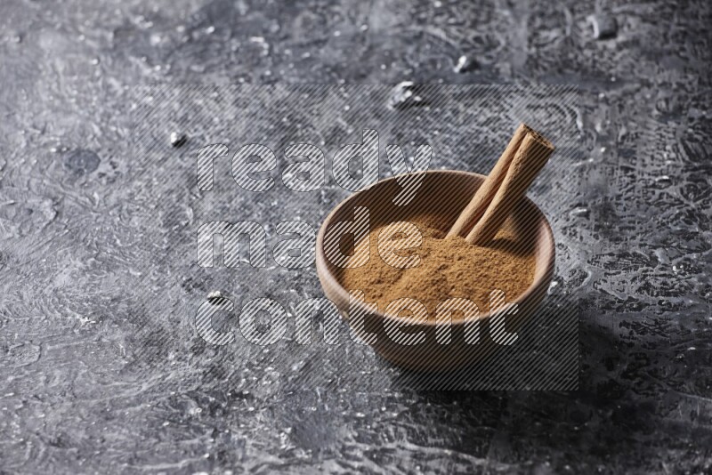 Wooden bowl full of cinnamon powder and a cinnamon stick on a textured black background