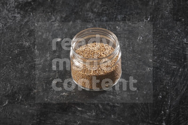 A glass jar full of mustard seeds on a textured black flooring