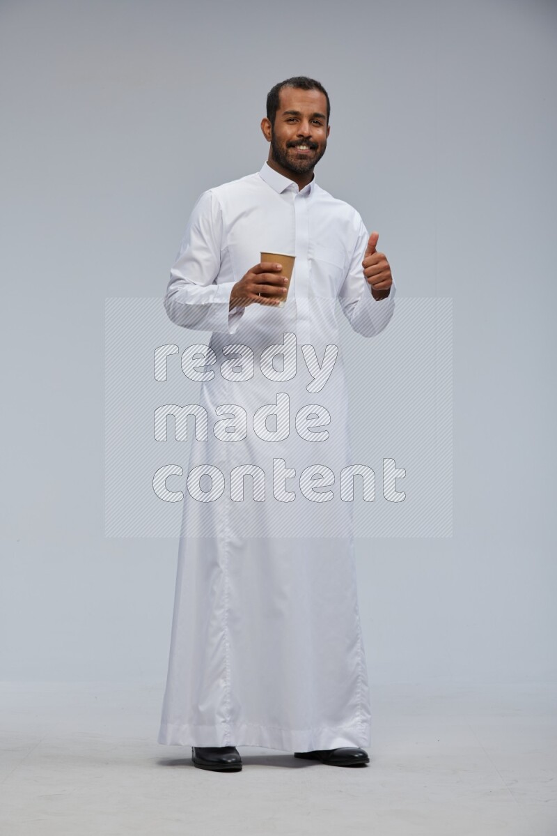 Saudi man wearing thob standing holding paper cup on gray background