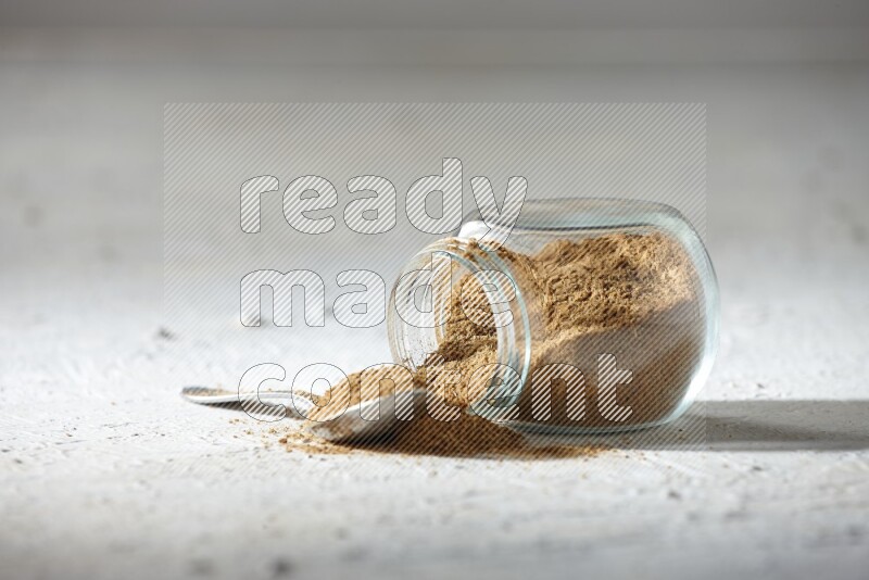 A flipped glass spice jar and a metal spoon full of cumin powder and powder spilled out on textured white flooring
