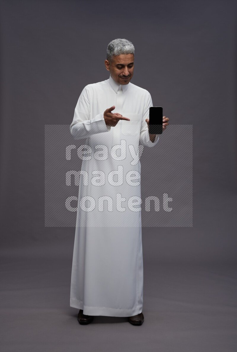 Saudi man wearing thob standing showing phone to camera on gray background