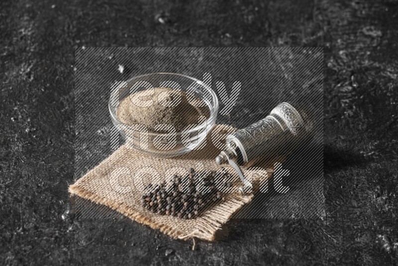 A glass bowl full of black pepper powder and black pepper beads on burlap fabric with a turkish metal pepper grinder on textured black flooring