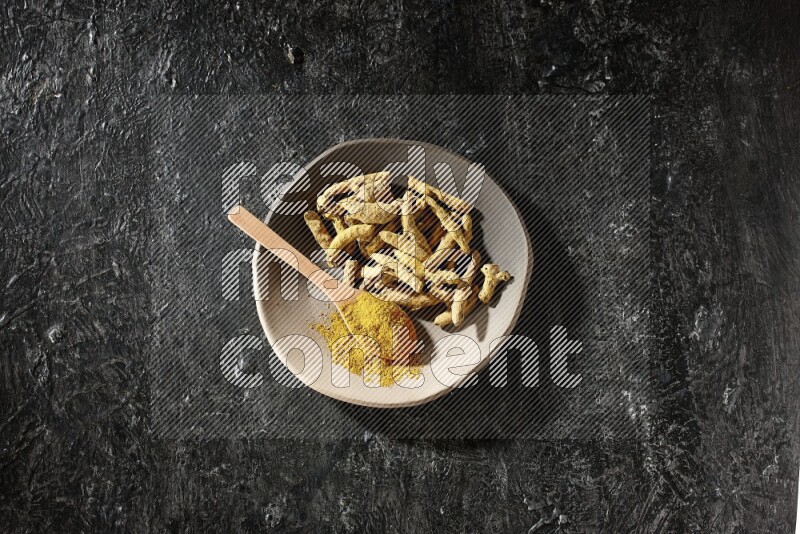A plate filled with dried turmeric fingers and a wooden spoon full of turmeric powder on a textured black flooring