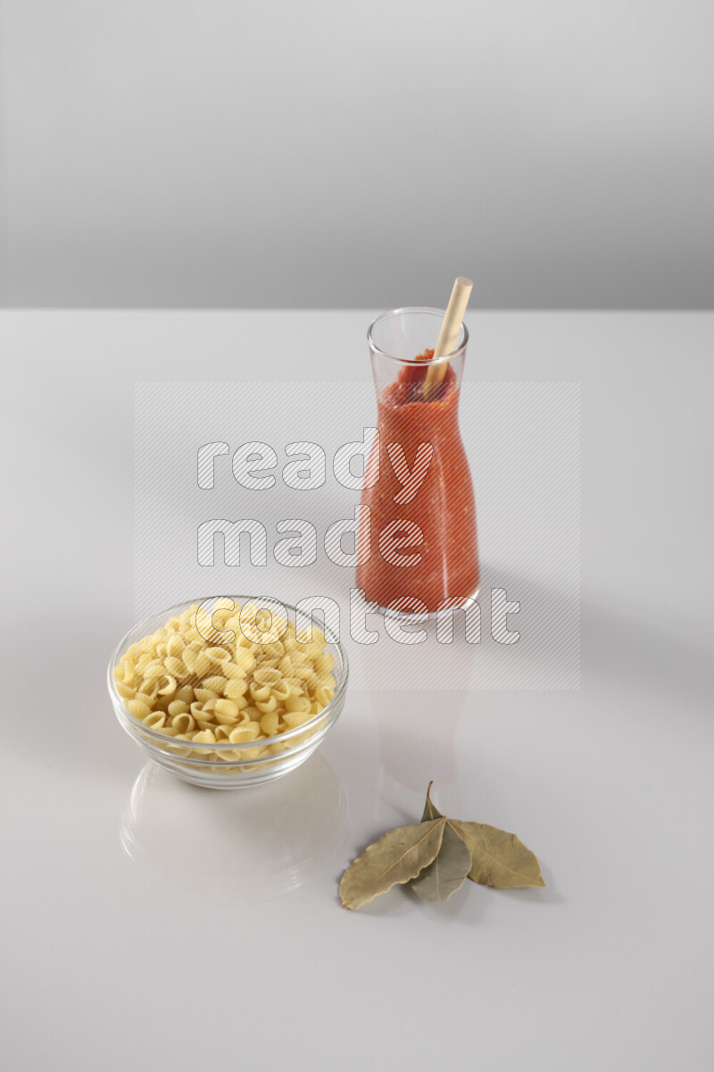 Raw pasta with tomatoe pasta with different ingredients such as cherry tomatoes, basil, garlic, bay laurel, cardamom, white pepper, black pepper, red chilis and wheat stalks on light grey background