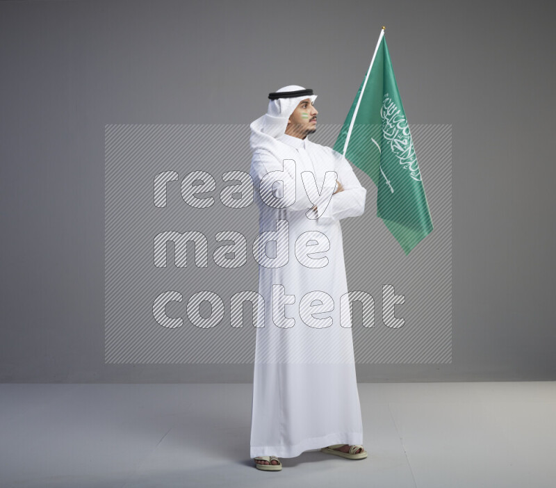 A Saudi man standing wearing thob and white shomag with face painting raising big Saudi flag on gray background