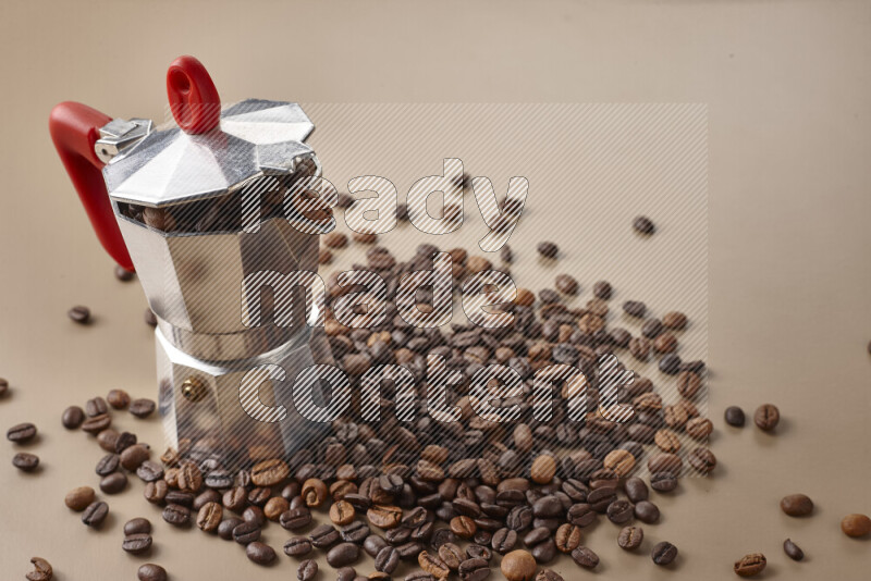 A moka pot with red handle surrounded by roasted coffee beans on beige background