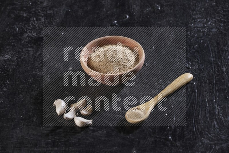 A wooden bowl and spoon full of garlic powder and beside it garlic cloves on a textured black flooring