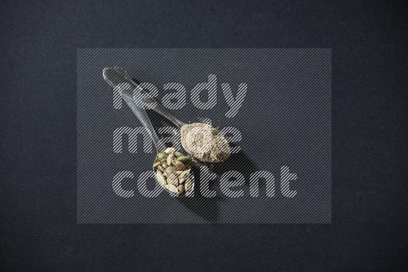 ملعقتان معدنيتان ممتلئتان ببودرة الهيل وحبوب الهيل على خلفية سوداء