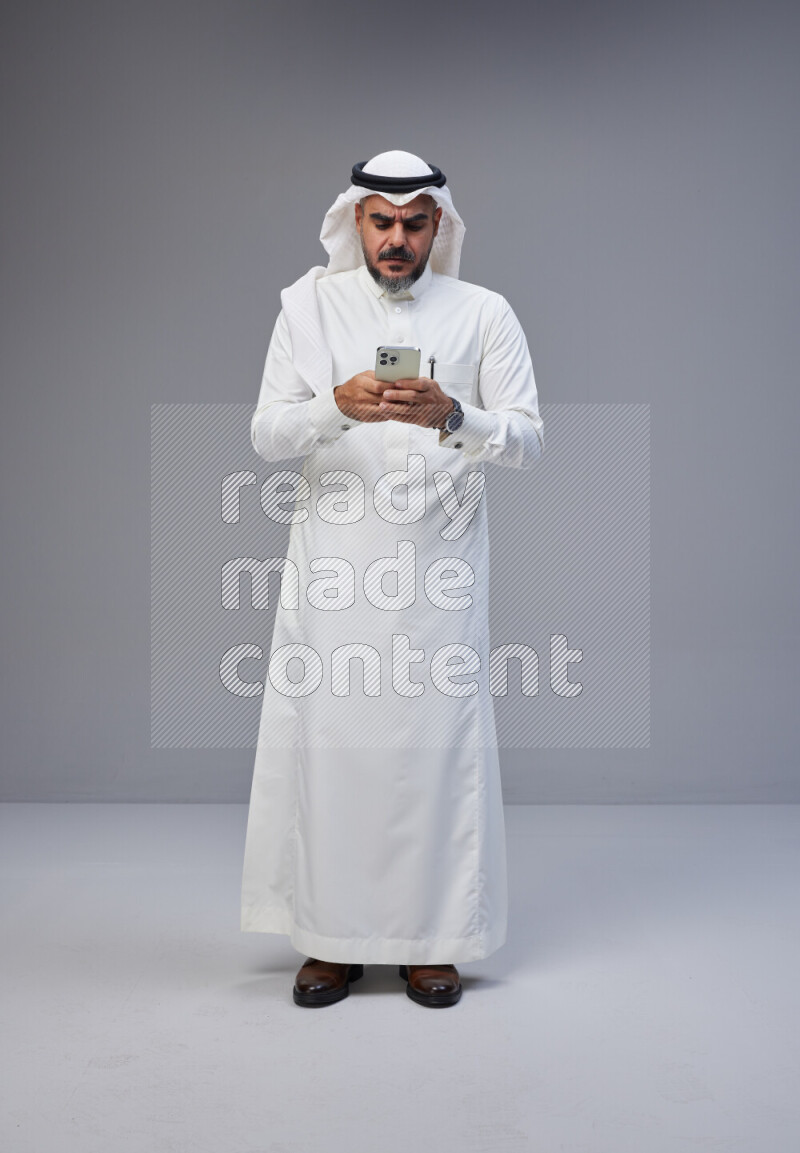 Saudi man Wearing Thob and white Shomag standing texting on phone on Gray background