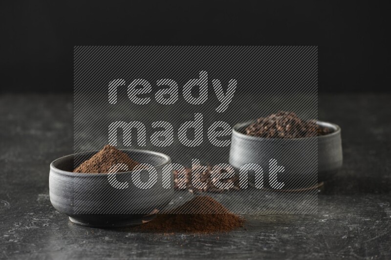 2 Black pottery bowls full of cloves and the other full of cloves powder on textured black flooring