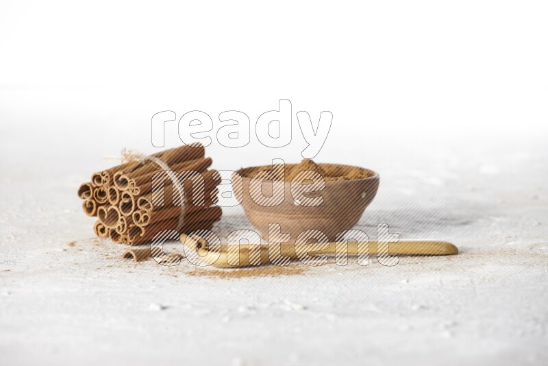 Cinnamon sticks stacked and bounded beside a wooden bowl full of cinnamon powder and a wooden spoon full of powder on white background