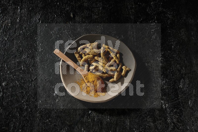 A plate filled with dried turmeric fingers and a wooden spoon full of turmeric powder on a textured black flooring
