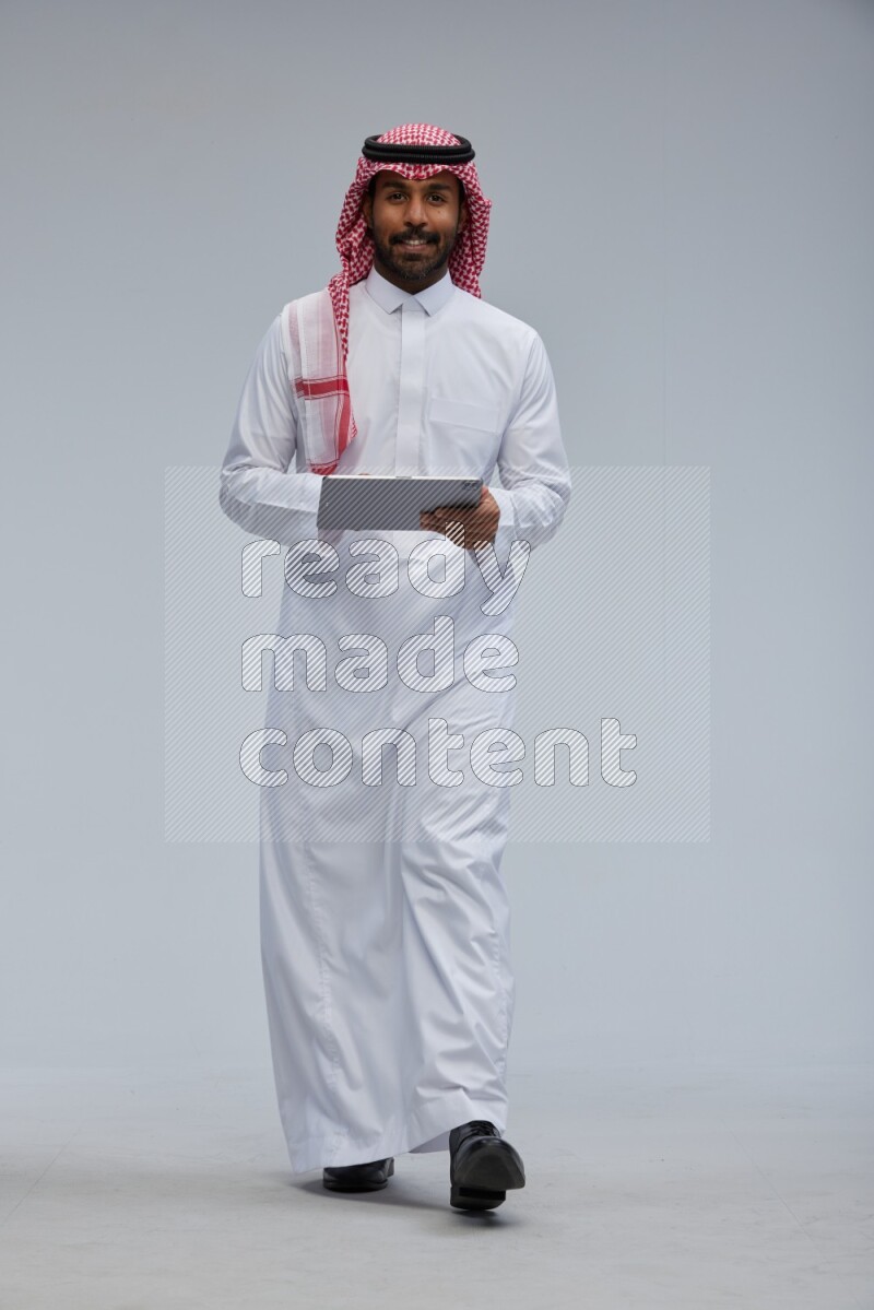 Saudi man Wearing Thob and shomag standing working on tablet on Gray background