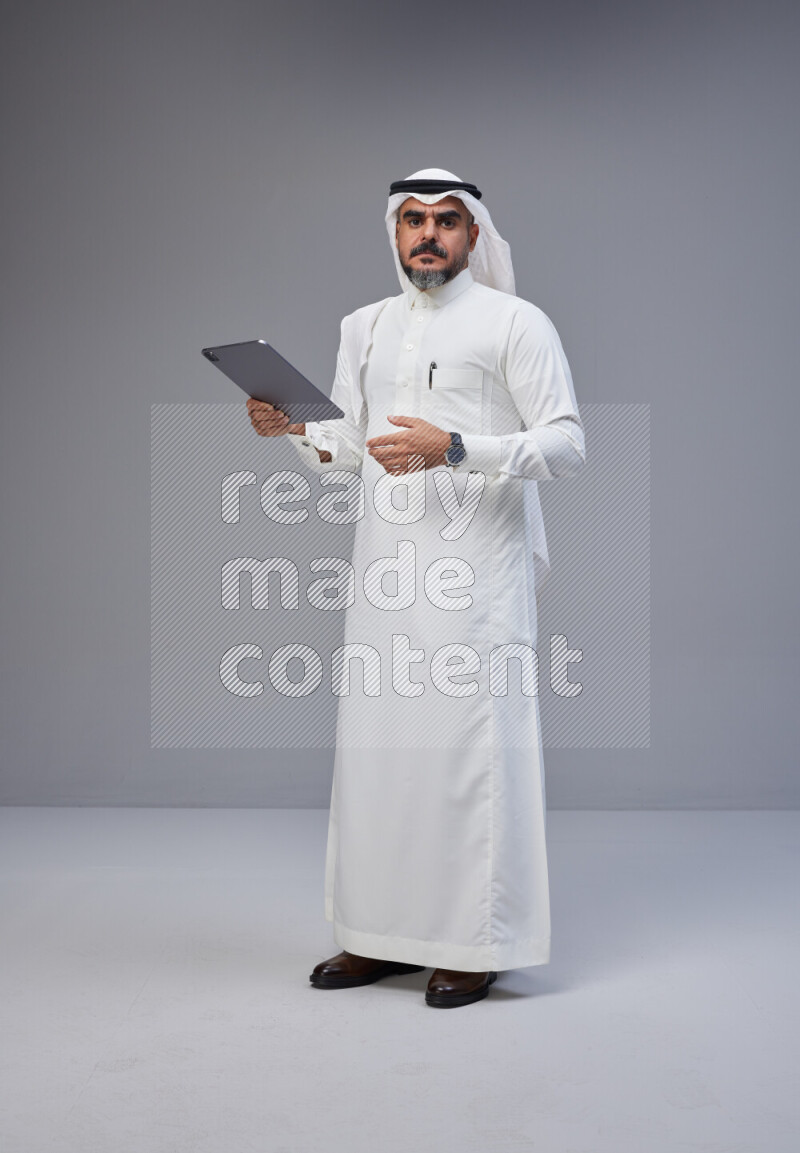 Saudi man Wearing Thob and white Shomag standing working on tablet on Gray background