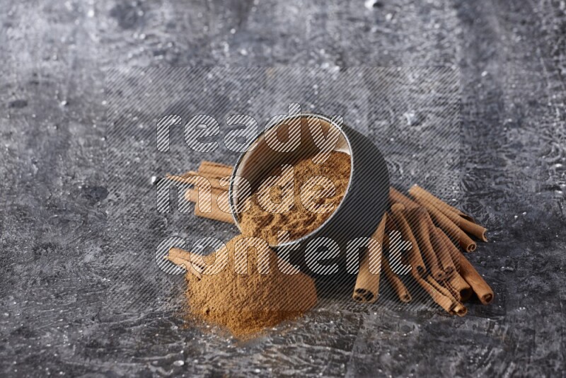 Black pottery bowl over filled with cinnamon powder and cinnamon sticks around the bowl on a textured black background