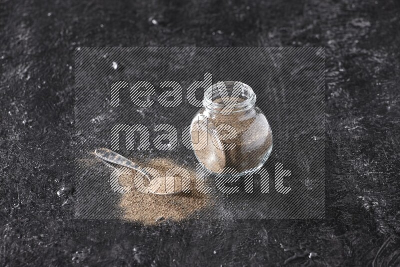 A glass spice jar full of black pepper powder and a metal spoon full of powder on textured black flooring