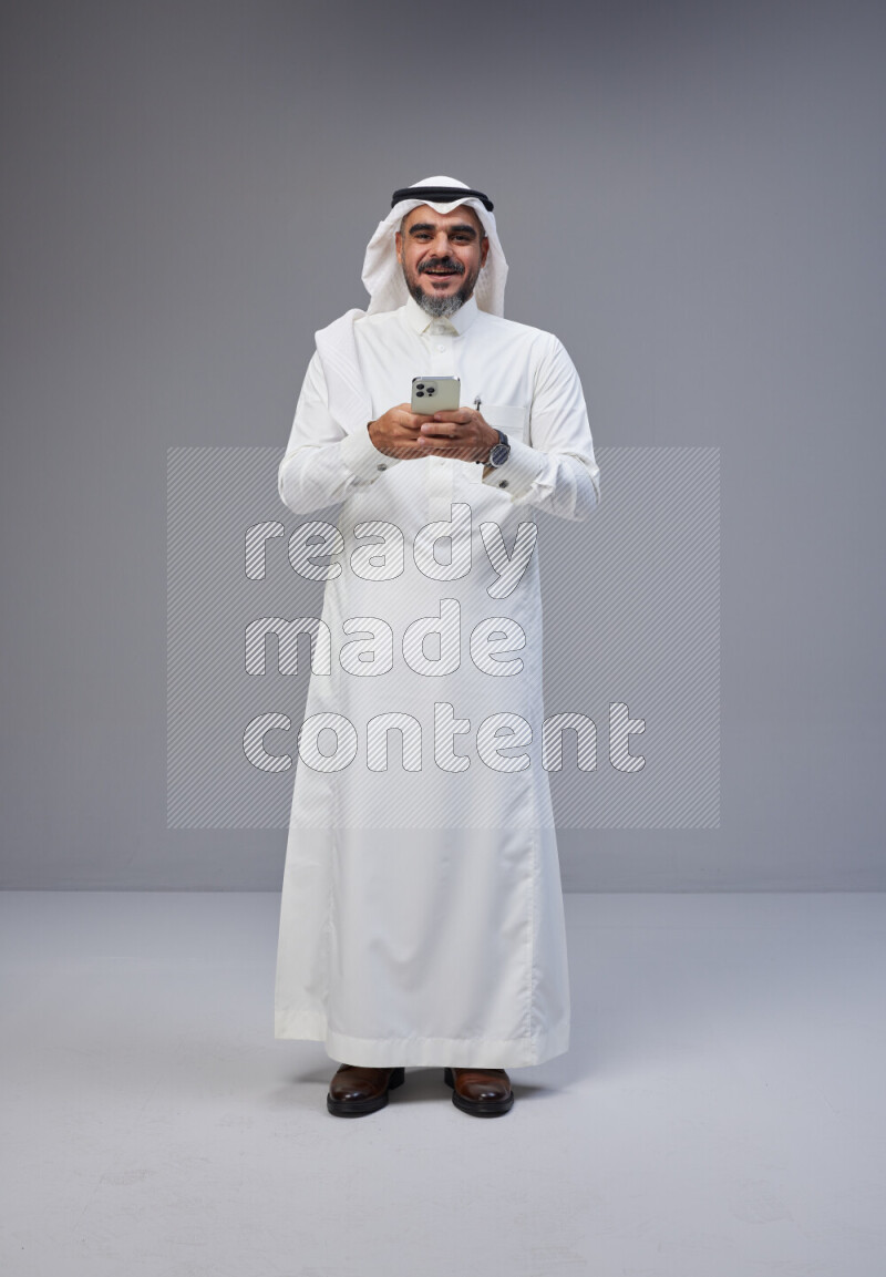Saudi man Wearing Thob and white Shomag standing texting on phone on Gray background