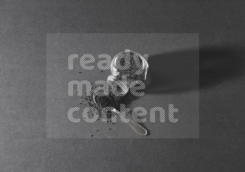 A flipped glass spice jar full of black seeds and the seeds spread out with a metal spoon full it on a black flooring