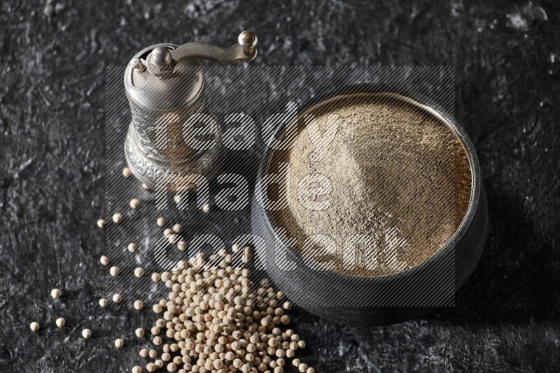 Black pottery bowl full of white pepper powder with pepper beads and metal pepper grinder on textured black flooring