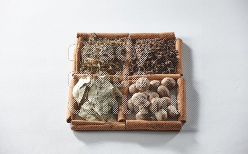 4 مربعات من أعواد القرفة ممتلئة بأوراق الغار والريحان المجفف والقرنفل وجوزة الطيب على أرضية بيضاء