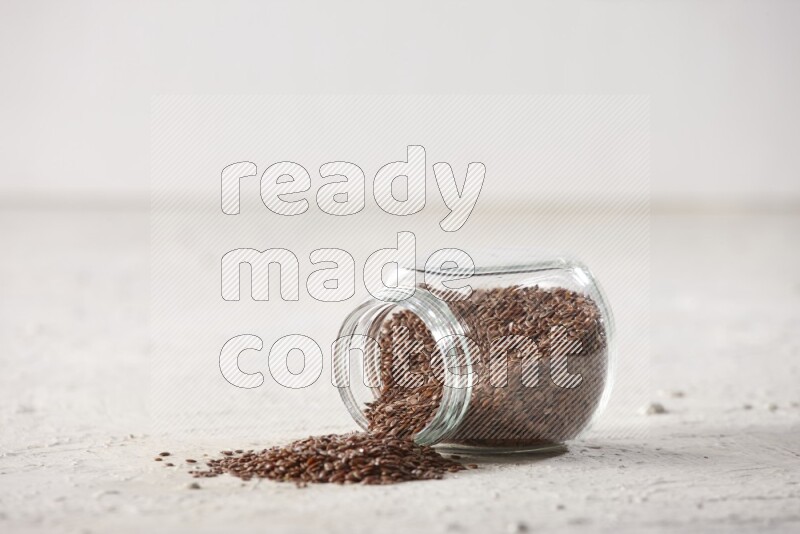 A glass spice jar full of flax seeds flipped and seeds spread out on a textured white flooring