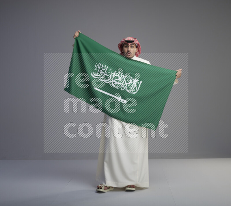 A saudi man standing wearing thob and red shomag holding big saudi flag on gray background