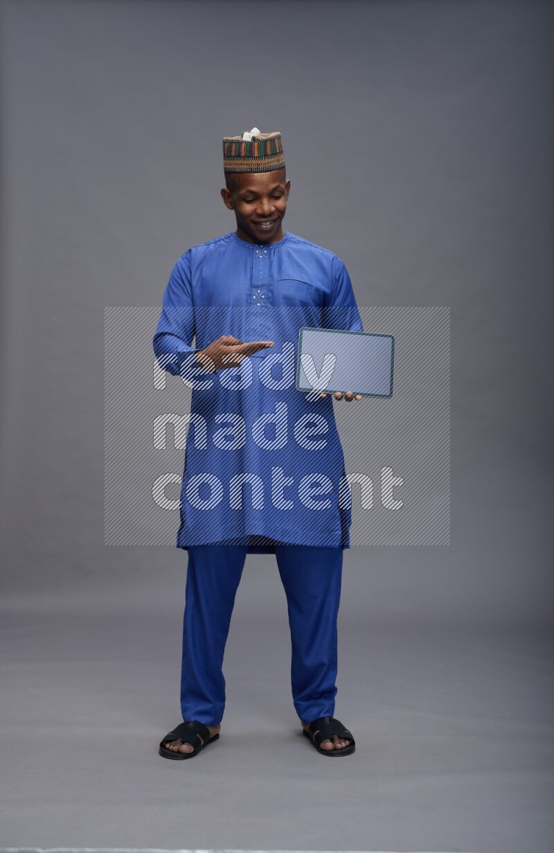 Man wearing Nigerian outfit standing showing tablet to camera on gray background