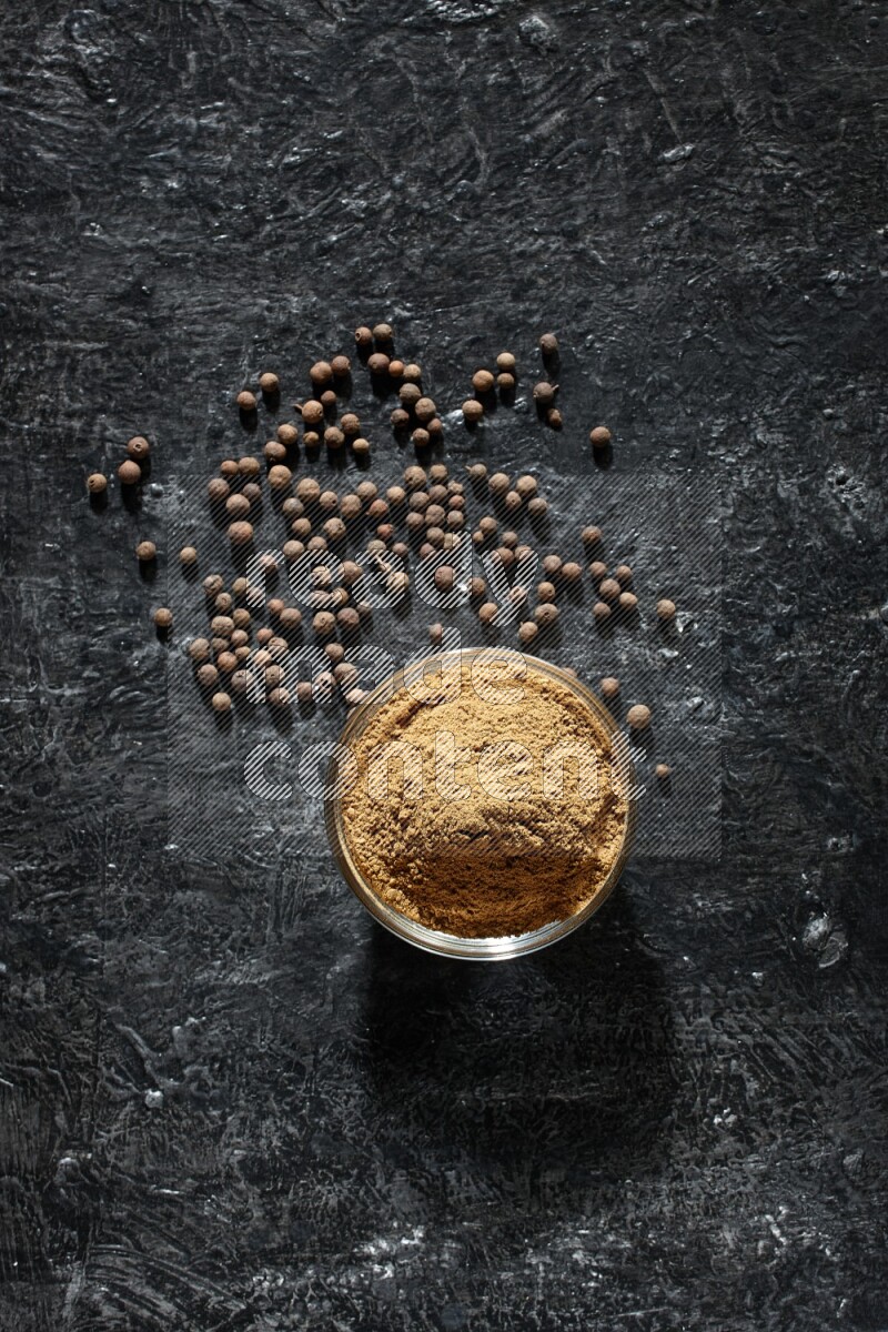 وعاء زجاجي ممتلئ ببودرة بهار الفلفل الحلو وحبوبه متناثرة على خلفية سوداء