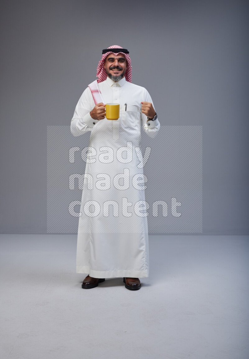 Saudi man Wearing Thob and red Shomag standing holding mug on Gray background