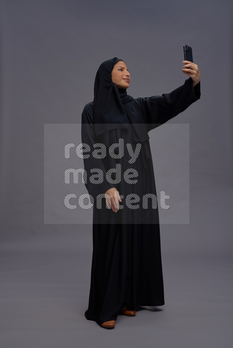 Saudi woman wearing Abaya standing taking selfie on gray background