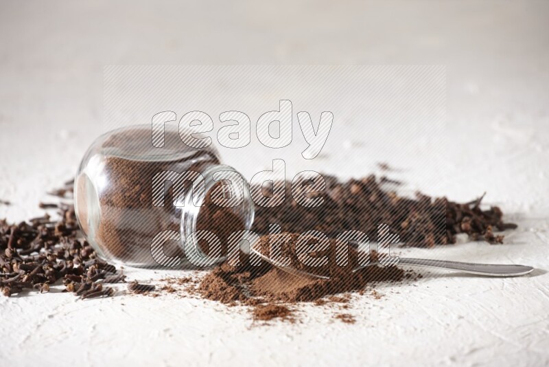 A flipped glass spice jar and a metal spoon full of cloves powder and powder came out of the jar with cloves spread on textured white flooring