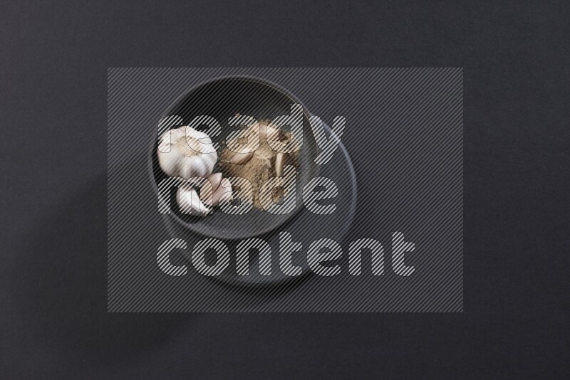 A black pottery bowl full of garlic powder, some cloves and a whole garlic bulb on a black flooring