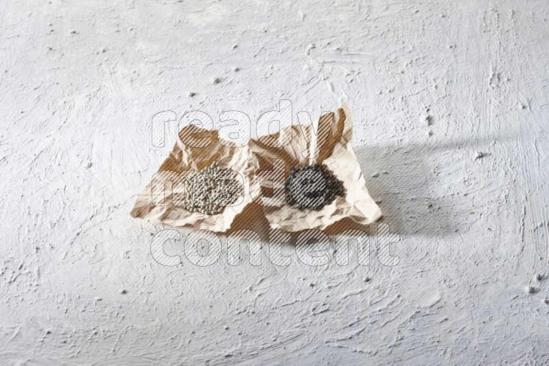 2 Crumpled pieces of paper full of black and white pepper beads on a textured white flooring