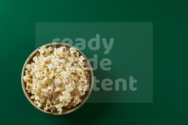 A beige ceramic bowl full of popcorn on a green background in a top view shot