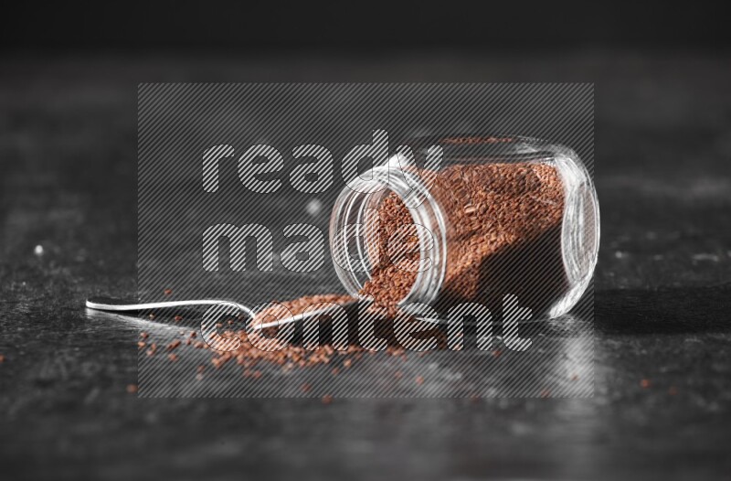 A glass spice jar full of garden cress seeds flipped and seeds spread out with a metal spoon full of the seeds on a textured black flooring