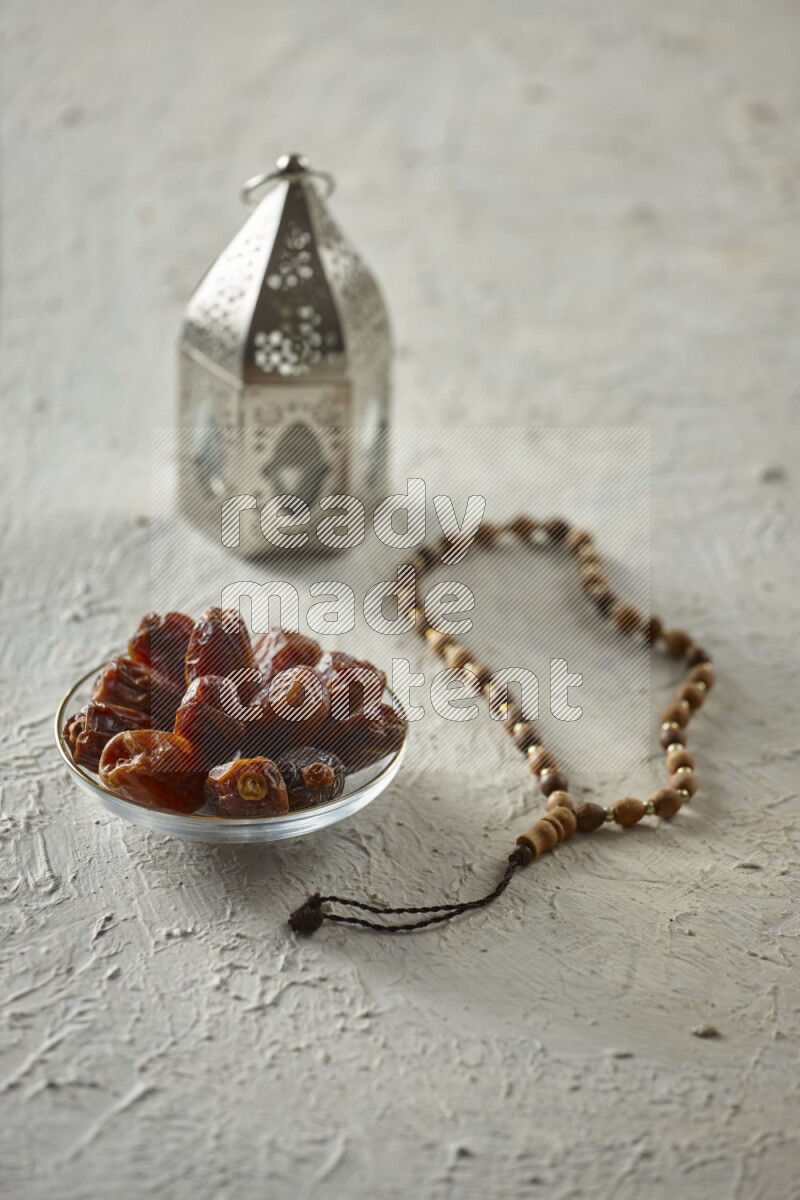 A silver lantern with different drinks, dates, nuts, prayer beads and quran on textured white background