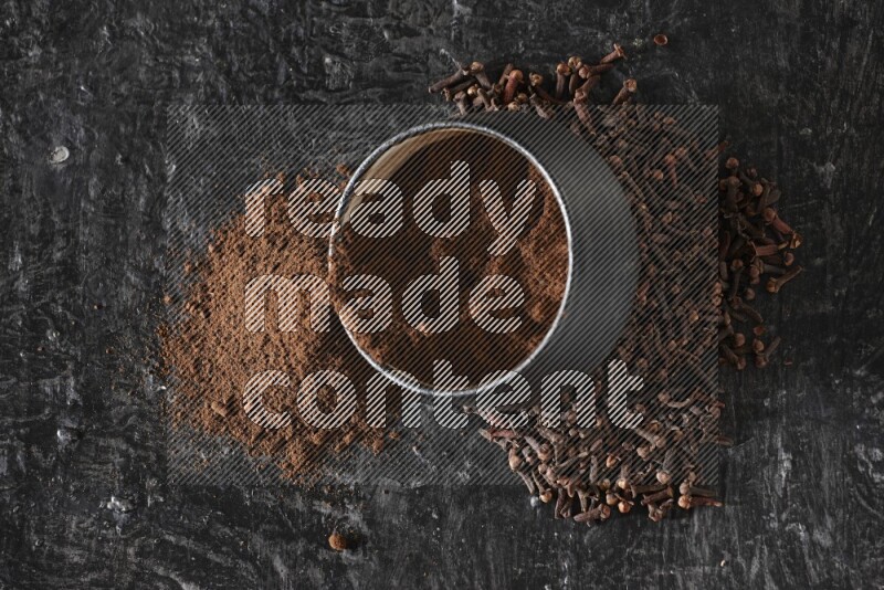 وعاء فخاري أسود ممتلئ ببودرة القرنفل والبودرة تناثرت منه مع الحبوب على خلفية سوداء