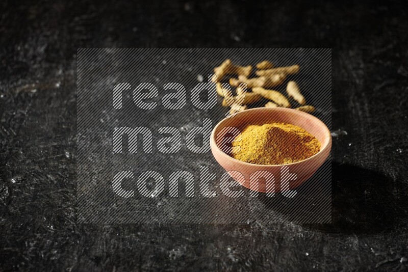 A wooden bowl full of turmeric powder with dried turmeric fingers on textured black flooring