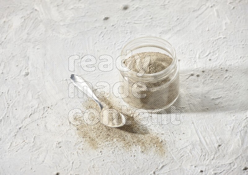 A glass jar and a metal spoon full of white pepper powder on textured white flooring