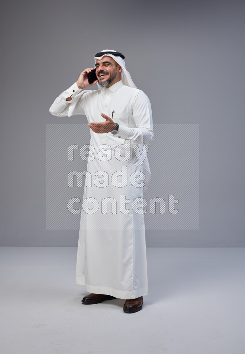 Saudi man Wearing Thob and white Shomag standing talking on phone on Gray background